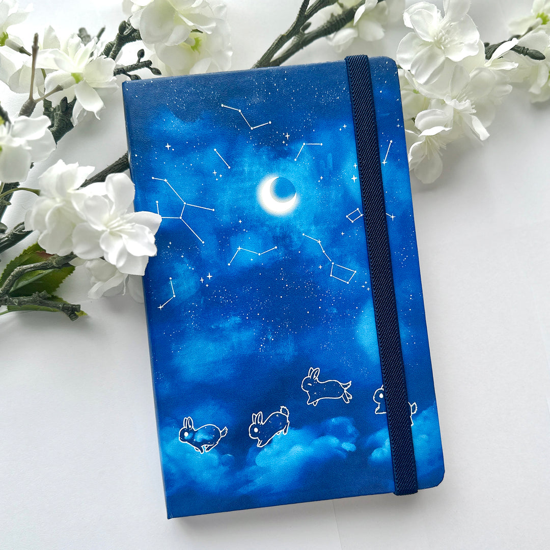 A notebook by Ika Ink. A5 sized leatherette cover notebook featuring original artwork of four cute little blue stellar bunnies dancing across the night sky while constellations and a crescent moon hand over head.